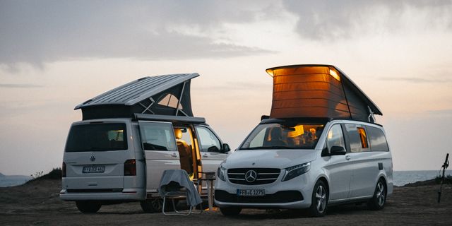 Zwei Camper mit aufgeklapptem Aufstelldach stehen beim Sonnenuntergang auf einem Hügel, innen brennt Licht.