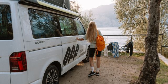 Frau neben VW Grand California Ocean auf Campingplatz vor See und Bergpanorama