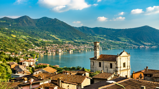 Oberitalienische Seen Geheimtipp: Blick auf eine Ortschaft am Iseosee in Norditalien  mit einem Kirchturm, dem Meer und Bergen vor dem blauen Himmel.