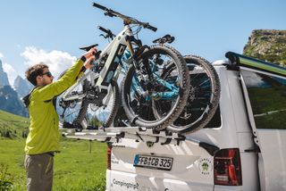 Ein Mann mit grüner Funktionsjacke und Sonnenbrille montiert während seinem Fahrradurlaub ein E-Bike auf dem Fahrradträger des CamperBoys Campers, der in einer Berglandschaft steht