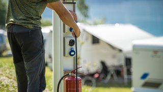 Landstrom Camper: Ein Mann schließt seinen Camper auf dem Campingplatz an den Landstrom an, im Hintergrund sieht man weiße Wohnmobile.