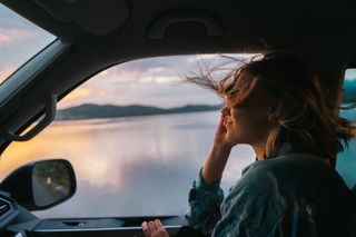 Eine Frau auf dem Beifahrersitz eines Campers. Sie lächelt und ihre Haare fliegen im Wind. Im Hintergrund der Sonnenuntergang.