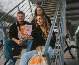 Three friends pose on stairs. They wear off merch: fanny pack, half zip sweater and jute bag