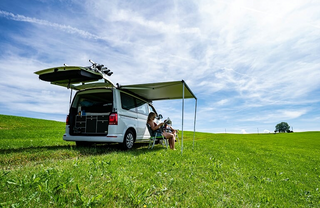 Eine Frau sitzt auf einer Wiese von einem CamperBoys Camper und liest Zeitung