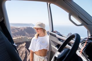 Eine Frau steht an der offenen Tür eines Campers. Hinter ihr erstreckt sich eine Küstenlandschaft.