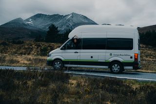 Ein VW Grand California fährt durch die schottische Landschaft. Eine Frau formt aus dem Fenster heraus mit den Händen ein Herz.