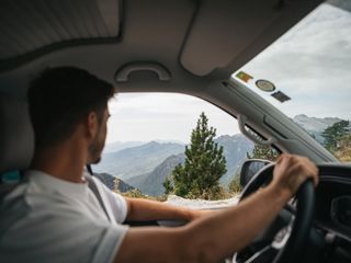 Ein Mann fährt einen Van beim Camping in Albanien und blickt durch das Fenster auf eine bewaldete Bergkulisse.