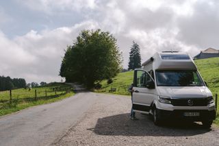 Ein Grand California Van steht in den Bergen geparkt. Ein Mann öffnet die Schiebetür. Auf einer Weide daneben grasen Kühe.