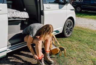 Eine Frau sitzt im offenen VW California Ocean und zieht sich Wanderschuhe an.