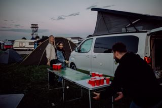 Zwei Frauen spielen eine Runde Bierpong gegen einen Mann, im Hintergrund sieht man ein Festivalgelände mit Zelten und einem Opel Crosscamp. Was du fürs Festival Camping alles brauchst, kannst du auf unserer Festival Packliste nachlesen.