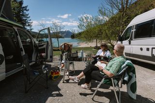 Drei Freunde beim Camping am See in Bayern sitzen an einem Campingtisch am Ufer