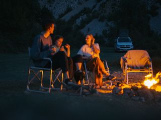 Camping Gadgets: Eine Freundesgruppe sitzt am Abend um ein Lagerfeuer herum, im Hintergrund sieht man ihren Camper.