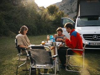 Drei Freund:innen frühstücken vor ihrem Off Camper auf der Wiese in einer Berglandschaft während die Sonne scheint.