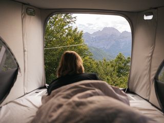 Eine junge Frau blickt aus dem Aufstelldach ihres Campers auf eine Berglandschaft beim Campen in den Dolomiten