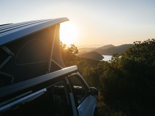 Das Aufstelldach eines Off Campers auf einem Berg, im Hintergrund sieht man Berge, Meer und die untergehende Sonne.