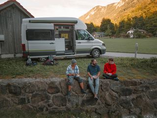 Drei Kletterfreunde machen Brotzeit. Im Hintergrund ist ein offener VW Grand California sowie Kletterequipment zu sehen.