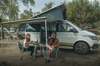 Ein Mann sitzen vor ihrem Camper an einem Campingtisch und trinken etwas.