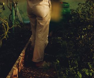 Frau in heller Hose barfuß zwischen Gemüsebeeten