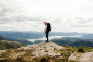 Ein Mann mit Wanderrucksack auf dem Rücken steht hoch oben auf einem Berg, im Hintergrund sieht man Berge und Wasser.