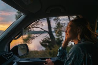 Eine Frau sitzt auf dem Beifahrersitz eines fahrenden Campers beim Überwintern in Portugal. Man sieht draußen den Sonnenuntergang.