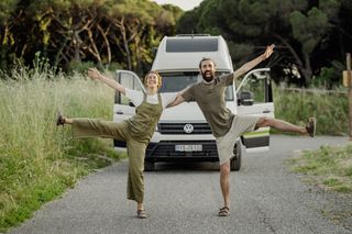 Ein Grand California Van ist auf einer ländlichen Straße geparkt. Zwei Menschen posieren davor und lachen.