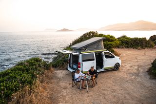 Camper an einem Strand mit zwei Menschen die am Tisch etwas essen