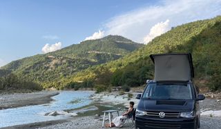 Julia, eine unserer Travel Designerinnen, sitzt vor einem Camper an einem Bach oder Fluss in den Bergen und lächelt in die Kamera.