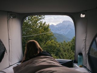 Eine Frau liegt in einem Camper im Bett und blickt auf eine bewaldete Berglandschaft.