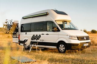 Ein Grand California 600 Camper steht auf einer Lichtung. Davor stehen Campingtische und es liegt eine Yogamatte auf dem Gras.